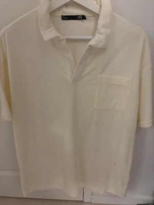 Krämvit henley med bröstficka - Krämvit henley  med klassisk krage och kort ärm. Skjortan har en enkel bröstficka och är tillverkad i mjuk bomull, perfekt för en clean och avslappnad look.