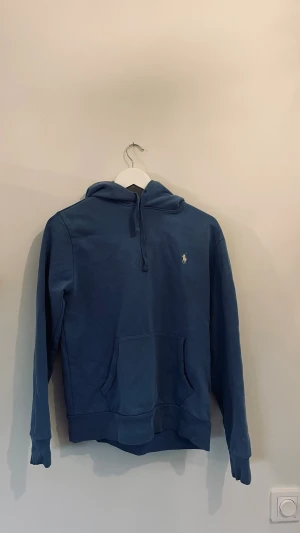 Blå hoodie från Polo Ralph Lauren XS/S - Snygg blå hoodie från Polo Ralph Lauren i storlek XS. Klassisk design med huva, snörning och magficka. Ikoniska broderade loggan på bröstet. Perfekt för dig som gillar stilrena och bekväma tröjor med premiumkänsla.