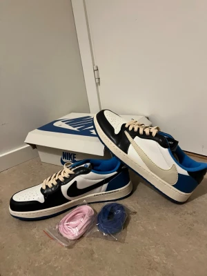 Nike Air Jordan 1 Low Travis Scott - Nike Air Jordan 1 Low Travis Scott x Fragment med svart, vit och blå färgblock, beige snörning och extra skosnören i rosa och blått. Skorna har ikoniska loggor på hälen och en platt blå sula. Tillverkade i skinn med perforerad tå och omvänd swoosh på sidan.
