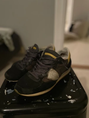Phillipe Model sneakers svart/gul - Säljer ett par Phillipe Model sneakers i svart med detaljer i gult och beige. Skorna har snörning, rund tå och platt sula. Materialmix av mocka och skinn med en beige panel på sidan och gul häl. Perfekt för dig som gillar exklusiva och unika sneakers. Trasig inuti vänster sko vid hälen (visas på sista bilden)