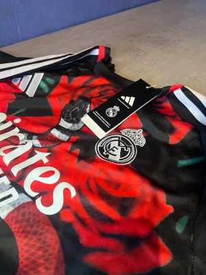 Real Madrid x Adidas röd/svart t-shirt - Säljer en Real Madrid t-shirt från Adidas med sjukt snyggt tryck av röda rosor och en stor orm över hela framsidan. Svarta och röda färger, vita Adidas-ränder på axlarna och Real Madrid-logga på bröstet. Materialet är lätt polyester, perfekt för fotboll.