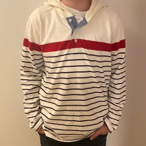 Randig hoodie från Hope & Henry XL - Vit hoodie med marinblå ränder och en bred röd rand över bröstet från Hope & Henry. Tröjan har huva, knappar vid halsen med blå detalj och är långärmad. Tillverkad i mjuk bomull, perfekt för en avslappnad stil.