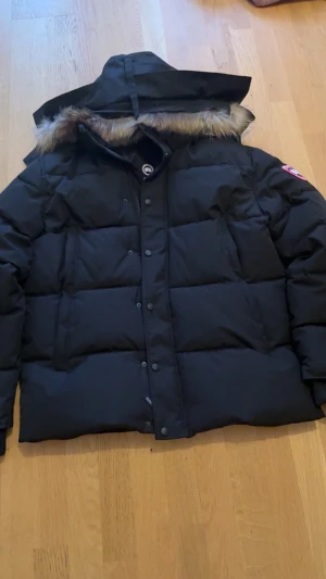 Svart dunjacka Canada Goose XL - Säljer en svart dunjacka från Canada Goose i storlek XL. Jackan har en fluffig huva med pälsdetalj, quiltad design och knappar framtill. Perfekt för kalla dagar med sin tjocka och varma fyllning. Klassisk logga på ärmen och innerfoder i svart.