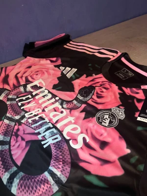 Real Madrid x Adidas rosa t-shirt L - Säljer en limited edition Real Madrid t-shirt från Adidas med sjukt snyggt rosa och svart blommigt mönster och en stor orm på framsidan. T-shirten har korta ärmar, klassiska Adidas-ränder på axlarna och är gjord i lätt polyester. Perfekt för dig som vill sticka ut på fotbollsplanen.