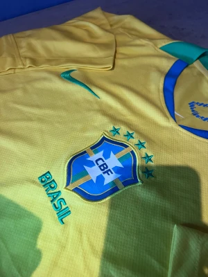 Brasilien Nike fotbollströja gul - Säljer en officiell Brasilien fotbollströja från Nike i klar gul med gröna och blå detaljer. Tröjan har korta ärmar, rund hals och broderat CBF-emblem på bröstet. Materialet är lätt polyester som andas, perfekt för match eller träning. Taggarna sitter kvar.