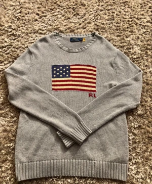 Grå stickad tröja Polo Ralph Lauren - Snygg grå stickad tröja från Polo Ralph Lauren med amerikansk flagga i blått, rött och vitt på bröstet samt RL-broderi. Jag har endast använt tröjan ett fåtal gånger då den är för stor för mig. Storlek på tröjan är S men den sitter som M eller till och med L. Den har rund hals och ribbade muddar vid ärmslut och nederkant. Perfekt för dig som vill ha en klassisk stil under vinter. Har du några frågor så är det bara att skriva👌