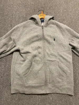 Grå hoodie från Polo Ralph Lauren - .Nu säljer jag denna populära polo Ralph lauren zipen. Skicket 8/10 💫. Storleken är 150-161cm men den är ganska liten i storleken. Passar mig som är 158cm. Denna Ralph lauren zipen är perfekt till sommarn🔥💫 hör av er vid funderingar🙏[ÄKTA]