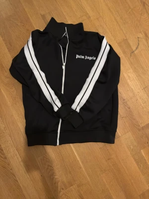 Palm Angels svart zip hoodie med vita ränder - Svart hoodie från Palm Angels med vita ränder längs ärmarna och vit dragkedja framtill. Tröjan har hög krage, ribbade muddar och logotypen broderad på bröstet. Materialet är mjuk polyester och passformen är normal. Perfekt för en sportig streetwear-look.