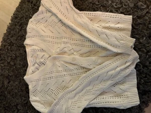 Vit stickad kofta från Lindex - Supersöt vit stickad kofta från Lindex med snyggt hålmönster och v-ringning. Koftan har knappar framtill och ribbade muddar vid ärmslut och nederkant. Perfekt att slänga över en topp eller en klänning när det blir lite kyligt.