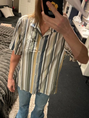 Randig skjorta från Bläck XL - Snygg skjorta från Bläck i storlek XL med vertikala ränder i vitt, blått, beige, grått och grönt. Skjortan har korta ärmar, klassisk krage och knäppning framtill. Tillverkad i mjuk bomull som känns skön mot huden. Perfekt för dig som gillar avslappnad stil.