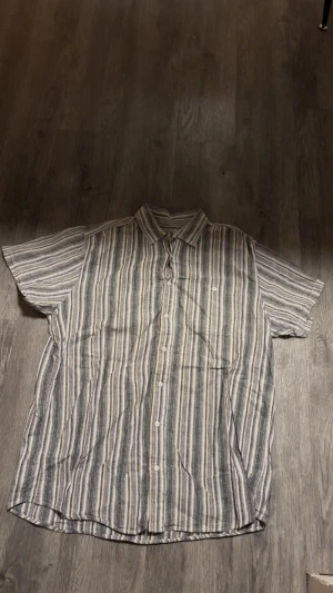 Randig kortärmad skjorta Dressmann XL - Kortärmad skjorta från Dressmann i storlek XL. Skjortan har ett randigt mönster i svart, grått och vitt, klassisk krage och knäppning framtill. Tillverkad i luftig bomull, perfekt för varma dagar. Snygg och avslappnad stil med bröstficka.