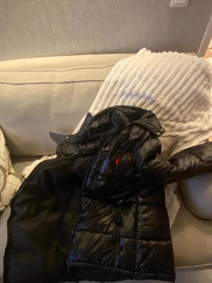 Svart pufferjacka från Polo Ralph Lauren - Svart pufferjacka från Polo Ralph Lauren med glansig finish och röd logga på bröstet. Jackan har hög krage, dragkedja framtill och quiltade paneler. Perfekt för kalla dagar och har en modern, loose passform.