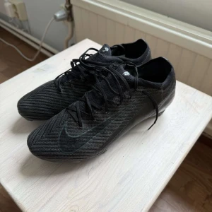 Svarta Nike Mercurial fotbollsskor - Säljer ett par svarta Nike Mercurial fotbollsskor med snyggt mönstrad ovandel och klassisk Nike-logga på sidan. Skorna har snörning och är tillverkade i syntetmaterial för bästa grepp och känsla på planen. Perfekta för dig som vill ha stilrena och snabba skor.