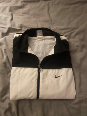 Svartvit vindjacka från Nike XL/M - Funkar om man har storlek M också.Nike vindjacka i svart och vitt med klassisk Swoosh-logga på bröstet. Jackan har hel dragkedja, ståkrage och meshfoder på insidan. Snygg sportig look med svarta ärmar och axelparti, samt vita detaljer framtill och baktill.