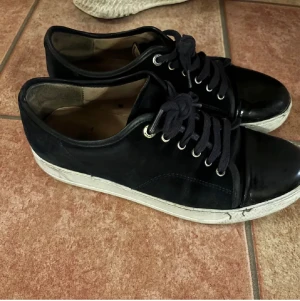 Lanvin sneakers med lackad tå - Snygga mörkblå sneakers med cool lackad tå och vita sulor. Skorna har svarta snören och silverfärgade öljetter. Ovandelen verkar vara i mocka eller syntet, vilket ger en schysst kontrast mot den blanka tån. Perfekta för dig som gillar en stilren men ändå unik look.