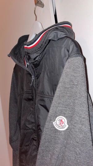 Moncler grå vind jacka /kofta  - Säljer en grå vindkofta från Moncler med stickade ärmar och luva. Koftan har dragkedja framtill, två fickor med dragkedja och Moncler-logga på ärmen. Snygga röd-vita detaljer vid luva och nederkant.  Koftan kan även användas som vindjacka. 