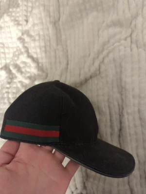 Svart Gucci keps med röd/grön rand - Säljer en svart keps från Gucci med klassisk röd och grön rand på sidan. Justerbar rem baktill i svart läder och metallspänne. Kepsen är tillverkad i Italien och har märkets logga på insidan. Perfekt för dig som gillar lyxiga streetwear-accessoarer.