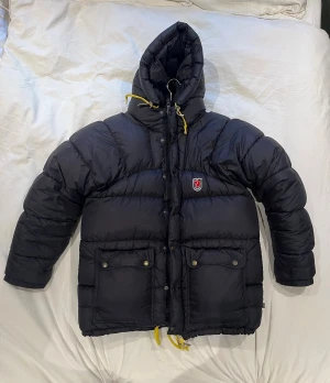Svart dunjacka från Fjällräven - SKCIKA PRISFÖRLAG!!                         Svart, puffig dunjacka från Fjällräven med gul snörning i huvan och nederkant. Jackan har två stora fickor med tryckknappar, dragkedja och Fjällräven-logga på bröstet. Perfekt för kalla dagar med sin rejäla huva och isolerande material. Liten lagning som syns på bild 6 samt att dragkedjan har tappat lite färg annars är den i väldigt fint skick! Gamla modellen dvs att den är lite större i modellen, nypris runt 10 000