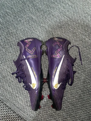 Lila Nike Mercurial fotbollsskor - Säljer ett par lila Nike Mercurial fotbollsskor med snygga guldiga och vita detaljer samt grafiska mönster i lila och guld. Skorna har snörning, platt sula med dobbar och är tillverkade i syntetmaterial för grym komfort och grepp på planen. Perfekt för dig som vill sticka ut på matchen!