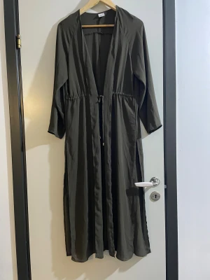 Mörkgrön långklänning med knyt - Snygg mörkgrön långklänning med knyt i midjan och långa ärmar. Klänningen har en avslappnad passform och är tillverkad i ett lätt, flowy material som ger en cool vibe. Perfekt för dig som gillar minimalistisk stil och vill ha något enkelt men ändå trendigt.