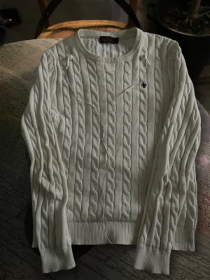 Stickad tröja från Morris - Snygg vit kabelstickad tröja från morris med rund hals och ribbade muddar. Tröjan har ett litet broderat morris-emblem på bröstet och är gjord i mjuk bomull. Perfekt för dig som gillar klassisk och clean stil.