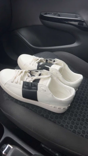 Valentino Open sneakers vit/svart - Säljer ett par snygga Valentino Open sneakers i vitt med en bred svart läderdetalj över snörningen. Skorna har platt sula, rund tå och coola nitar på hälen. Perfekt för dig som gillar stilrena och lyxiga sneakers med unik design.VILL FÅ SÅLT!