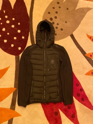 Svart pufferjacka från Lavetti Canada - Svart pufferjacka från Lavetti Canada med huva och snygga detaljer på axlarna. Jackan har quiltad framsida, dragkedja och ett rundat märke med röd lönnlöv på bröstet. Ärmarna är i ett mesh-liknande material för extra komfort och stil.