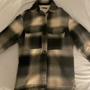 Rutig overshirt  - Snygg overshirt från i svart, grått och beige rutigt mönster. Insidan är fodrad med mjuk fuskpäls som gör den extra skön och varm. Modellen har klassisk krage, två bröstfickor och knappar framtill. Perfekt för lager-på-lager och chill stil.