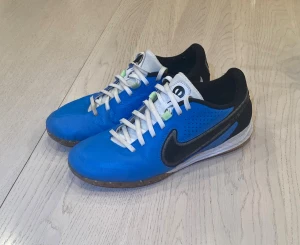 Nike Tiempo blå fotbollsskor - Säljer ett par Nike Tiempo fotbollsskor i blått med svarta och vita detaljer. Skorna har en låg profil, rund tå och platt sula med texten 'TIEMPO' under. Ovandelen är i läder med snörning och perforerade sidor för extra ventilation. Perfekt för inomhusspel.