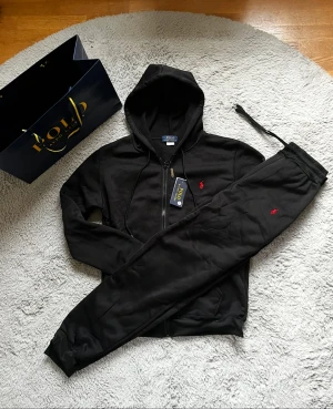 Ralph Lauren hoodie och byxa - Säljer ett Ralph Lauren kit i svart med röd broderad på bröst och vänstra benet. Storlekarna på både plaggen är s och mått finns att se i några av bilderna. Kläderna är helt nya och har prislapp kvar. 📦Skickas snabbt och paketeras med omsorg. OBS! Påsen som visas i några av bilderna används bara som dekoration och ingår inte i köpet.