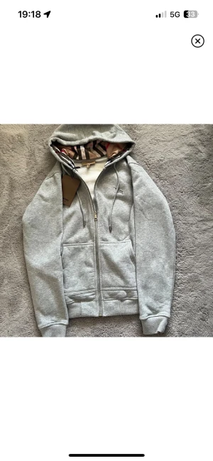 Grå Burberry hoodie med dragkedja - Snygg grå hoodie från Burberry med klassiskt rutigt foder i huvan och insidan. Tröjan har broderad logga på bröstet, snörning i huvan och praktiska fickor framtill. Materialet är mjuk bomull och polyester, perfekt för chill dagar.