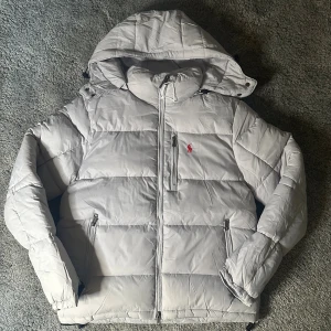 Ljusgrå pufferjacka från Polo Ralph Lauren - Säljer en ljusgrå pufferjacka från Polo Ralph Lauren med huva och dragkedja framtill. Jackan har två sidofickor med dragkedja och den klassiska röda Polo-loggan broderad på bröstet. Perfekt för kalla dagar och har en clean, modern look.