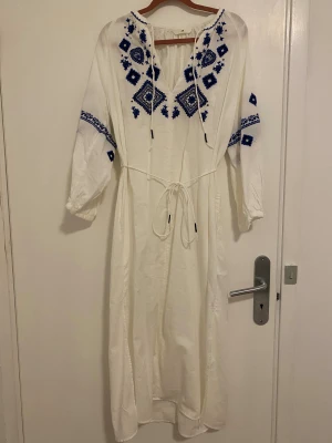 Vit långklänning med blå broderier H&M - Lång, luftig vit klänning från H&M med blå broderade blommor och geometriska mönster på bröst och ärmar. V-ringad med snörning i halsen och midjeband för snygg passform. Långärmad och med slits nertill. Perfekt för sommarens alla tillfällen.