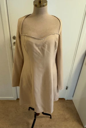 Beige kort klänning från Missguided - Snygg beige kortklänning från Missguided med långa ärmar och öppen rygg. Klänningen har en unik skärning vid axlarna och pärldetalj i nacken. Tillverkad i ett mjukt polyestertyg som ger en clean och stilren look.