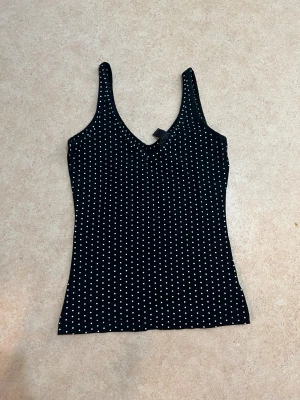 Svart prickig linnetopp i bomull - Svart linnetopp med vita prickar, djup v-ringning och smala axelband. Toppen är figurnära och tillverkad i mjuk bomull, perfekt för dig som gillar enkel men snygg stil. Storlek EUR 36-38. Tillverkad i Litauen.