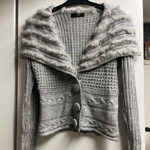 Grå kofta med pälsdetaljer i ullmix - Supermysig grå kofta med fluffiga pälsdetaljer på den breda kragen. Stickad i olika strukturer med flätmönster och stora knappar framtill. Långärmad, storlek S, normal i storleken. Tillverkad i en mix av ull, bomull och akryl. Mycket bra skick.