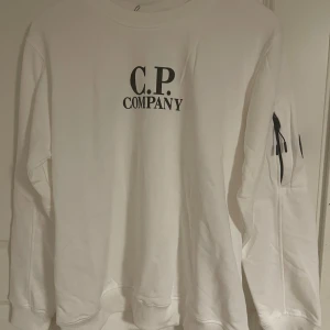 Vit sweatshirt från C.P. Company - Vit sweatshirt från C.P. Company med svart logotyp på bröstet och en unik svart linsdetalj på ärmen. Tröjan har rund hals, ribbade muddar och en dragkedja på ärmen. Perfekt för dig som gillar streetwear och snygga detaljer.
