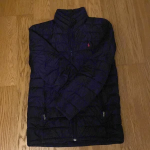 Mörlblå pufferjacka från Polo Ralph Lauren - Mörkblå pufferjacka från Polo Ralph Lauren med klassisk röd logga broderad på bröstet. Jackan har ståkrage, dragkedja framtill och två sidofickor med dragkedja. Quiltad design och lätt vadderad för extra värme. Perfekt för kyliga dagar. Sitter som s och inte använd mycket. 
