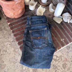 Blå Nudie Jeans, storlek 28/32 - Snygga blå jeans från Nudie Jeans med klassisk orange söm på bakfickorna. Modellen har slim/ raka ben och slitningar på framsidan för en cool, använd look. Riktigt skön wash och feta slitningar vid knäna