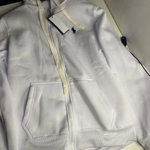 Vit hoodie från Polo Ralph Lauren - Vit hoodie från Polo Ralph Lauren med broderad svart logga på bröstet. Slim fit-modell med dragkedja, huva och ribbade muddar. Utrustad med två fickor framtill och snörning i huvan. Materialet är mjuk bomull med stretch för extra komfort.