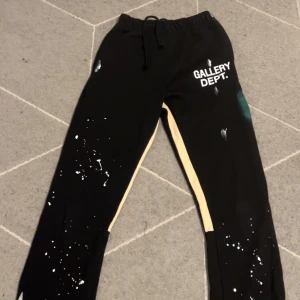 Svarta Gallery Dept mjukisbyxor S - Svarta mjukisbyxor från Gallery Dept med vit logga på höften, snörning i midjan och beige sidopaneler. Byxorna har coola vita färgstänk och en avslappnad passform. Perfekta för dig som gillar streetwear och unik design.