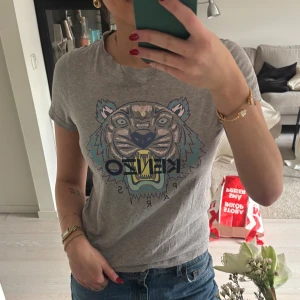 Grå Kenzo t-shirt med tigertryck - Säljer en grå t-shirt från Kenzo med ikoniskt tigertryck och Kenzo Paris-logga framtill. T-shirten är i mjuk bomull, har rund hals och korta ärmar. Perfekt för dig som gillar streetstyle och vill sticka ut med ett coolt motiv.