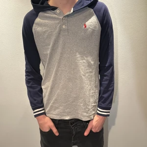 Grå-blå hoodie från Ralph Lauren - Snygg hoodie från Ralph Lauren med grå kropp och marinblå ärmar. Tröjan har huva, knappar vid halsen och en broderad röd logga på bröstet. På ena ärmen finns en stor vit 3:a. Muddar med vita ränder. Perfekt för en chill och sportig stil. Modellen är 186 cm och väger 70 kg 