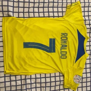Al Nassr Ronaldo fotbollströja Nike - Säljer en gul Al Nassr fotbollströja med blå detaljer, tryck med Ronaldo och nummer 7 på ryggen. Framsidan har klubbmärke, sponsor KAFD och Nike-logga. Kortärmad modell i lätt, ventilerande material som passar perfekt för match eller träning.