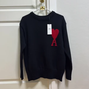 Svart stickad tröja från Ami - Svart stickad tröja från Ami med ett stort rött hjärta och bokstaven A broderat på bröstet. Rund hals och långa ärmar med ribbade muddar. Perfekt för dig som gillar stilrena detaljer och vill ha något som sticker ut.