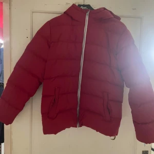 Röd dunjacka från United Colors of Benetton - Snygg röd dunjacka från United Colors of Benetton med huva och dragkedja framtill. Jackan har ett tryck på huvan och är perfekt för kyligare dagar. Den är vadderad och ger en skön värme.