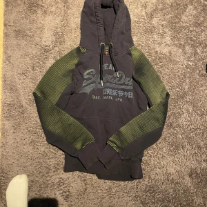Svart Superdry hoodie med gröna detaljer - Cool svart hoodie från Superdry med gröna, tunna ränder på ärmarna. Stor logga och japansk text framtill, justerbar huva med snörning och känguruficka. Perfekt för dig som gillar streetwear och vill sticka ut lite extra.