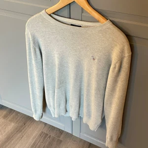 Ljusgrå stickad tröja från Gant - Snygg ljusgrå stickad tröja från Gant med rund halsringning och diskret broderad logga på bröstet. Tröjan har ribbade muddar vid ärmslut och nederkant, perfekt för en clean och klassisk stil.
