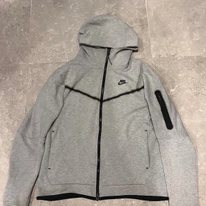 Grå nike tech - Grå vindjacka från Nike med svart dragkedja och huva. Jackan har en stilren design med svarta detaljer, inklusive en ficka med dragkedja på ärmen. Perfekt för dig som gillar sportig och clean look.