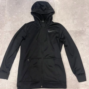Svart vindjacka från Nike med huva - Svart Nike vindjacka med huva och dragkedja hela vägen. Jackan har två fickor med dragkedja, långa ärmar och en stilren design med Nike-logga på bröstet. Perfekt för dig som gillar sportig och clean stil.